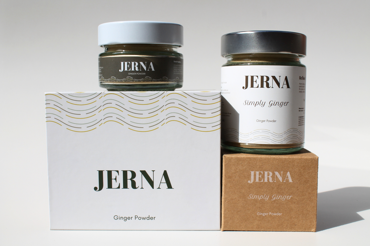 Two jars of JERNA Simply Ginger ginger powder displayed with their packaging boxes on a clean white background. One jar has a white lid and sits on a white box with gold wave details, while the other has a silver lid and sits on a brown kraft box.